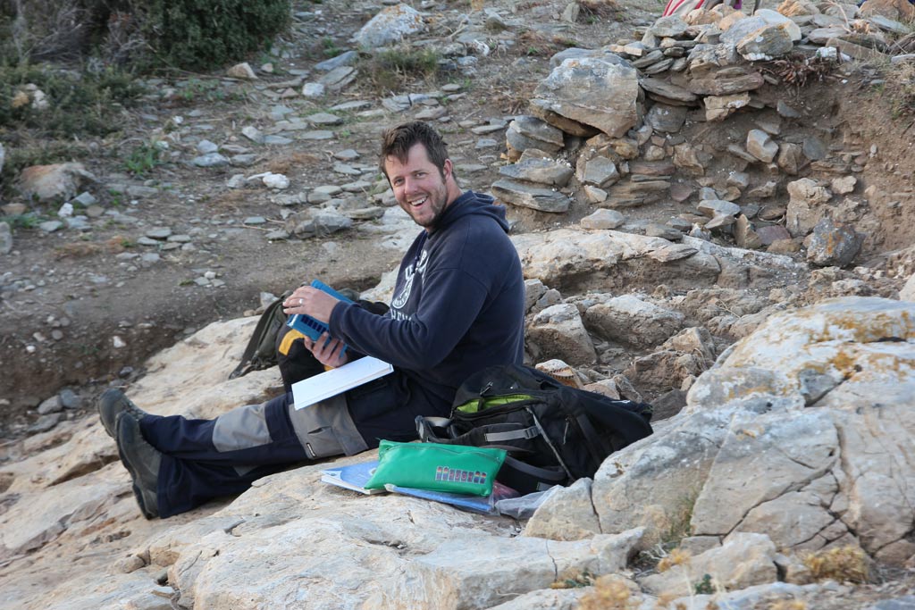 Hugh Thomas sitting on bedrock in Excavation Area 3 examining the Munsell Chart for information about soil colour