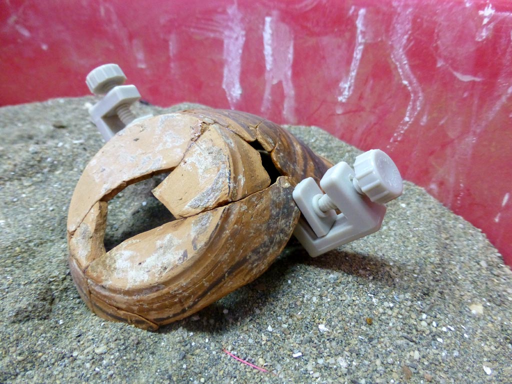 Pieces of a small fineware vessel held together with clamps and supported in sand to help the adhesive to gain its full strength